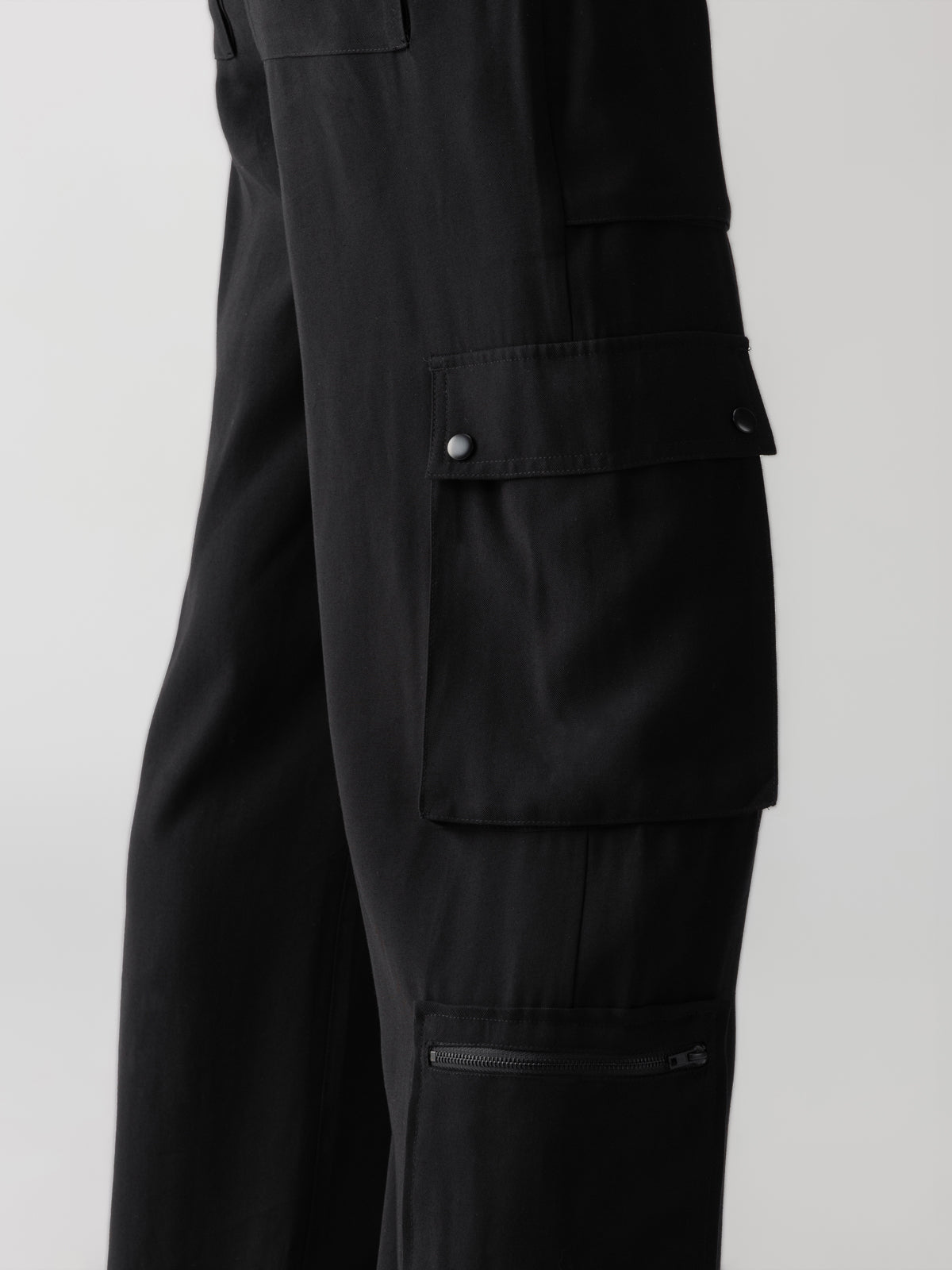 Close-up of the side view of the Ott Cargo Black pants by Sanctuary Clothing. Visible details include a large flap pocket with snap buttons on the thigh and a smaller zippered pocket below it. The fabric appears sturdy, suitable for both casual and functional use. The background is plain white.