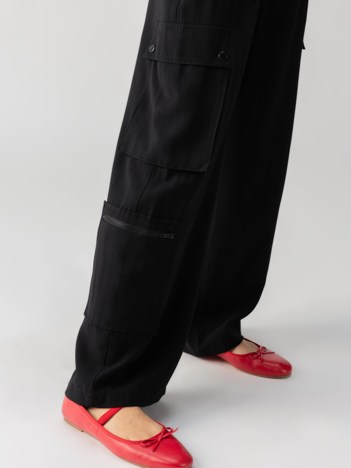 A person wearing Ott Cargo Black pants by Sanctuary Clothing and red ballet flats stands on a light gray surface. The pants feature multiple pockets, including a zipped one, and the flats have a simple design with a small bow on each shoe. Only the lower half of the person's body is visible.