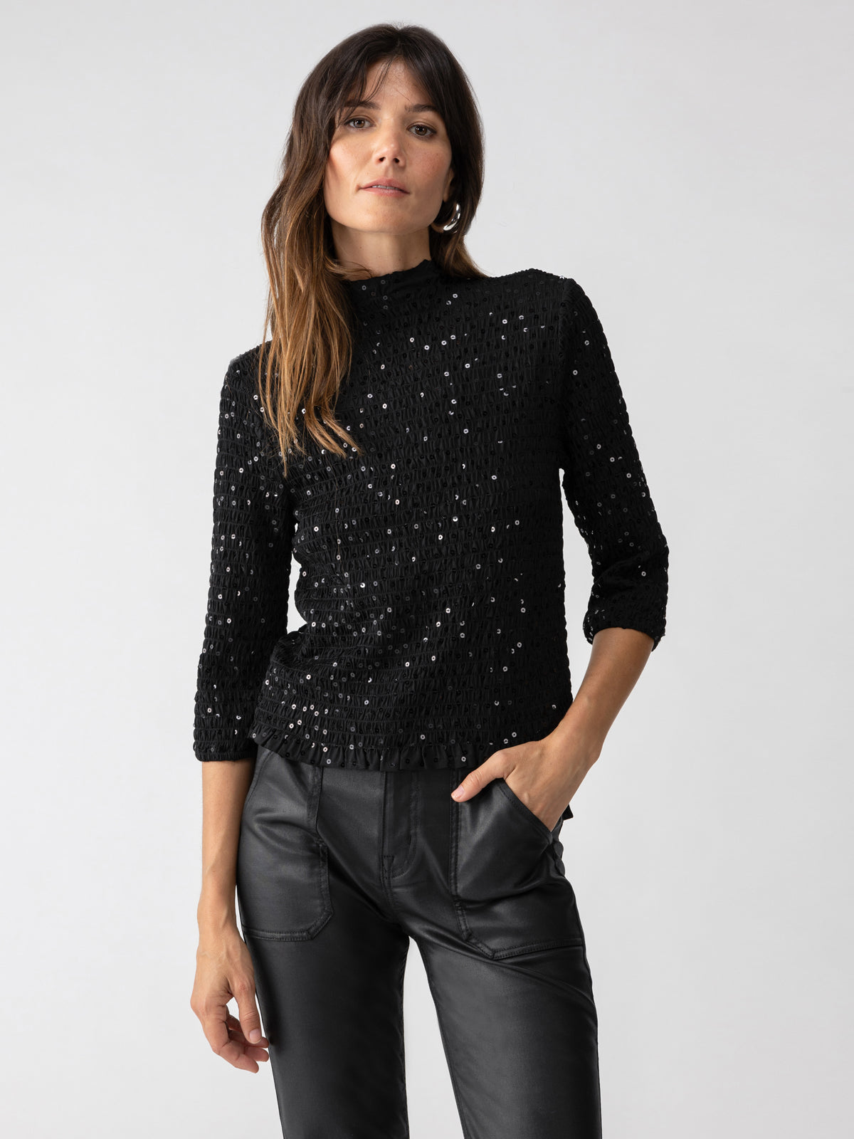 A woman standing against a plain background, wearing the "Almost Holiday Black" long-sleeved top with a sparkly texture and black leather pants by Sanctuary Clothing. Her left hand is in her pocket, and she has long brown hair styled with gentle waves.