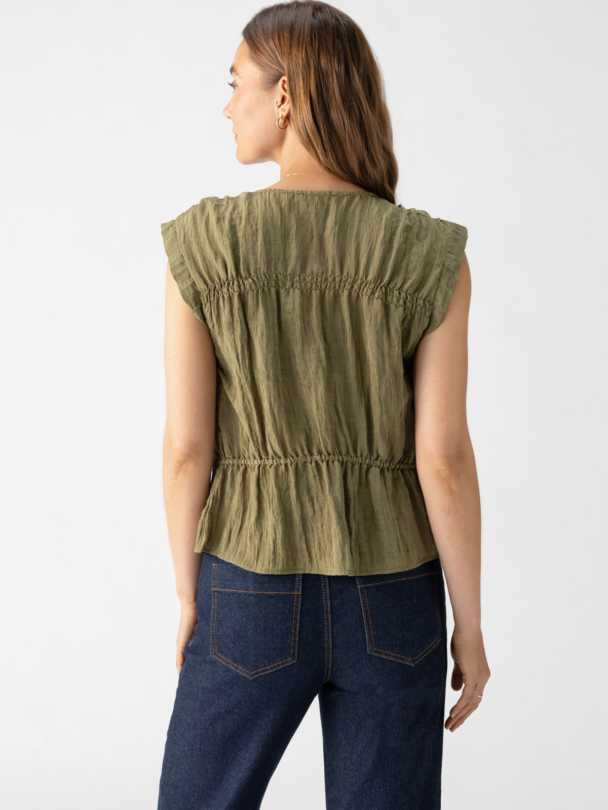 A woman with long brown hair is seen from the back, wearing the Gathered Shell Burnt Olive blouse by Sanctuary Clothing, featuring ruffled sleeves and cinched detailing at the waist. She pairs the top with dark blue jeans against a plain, light-colored background.