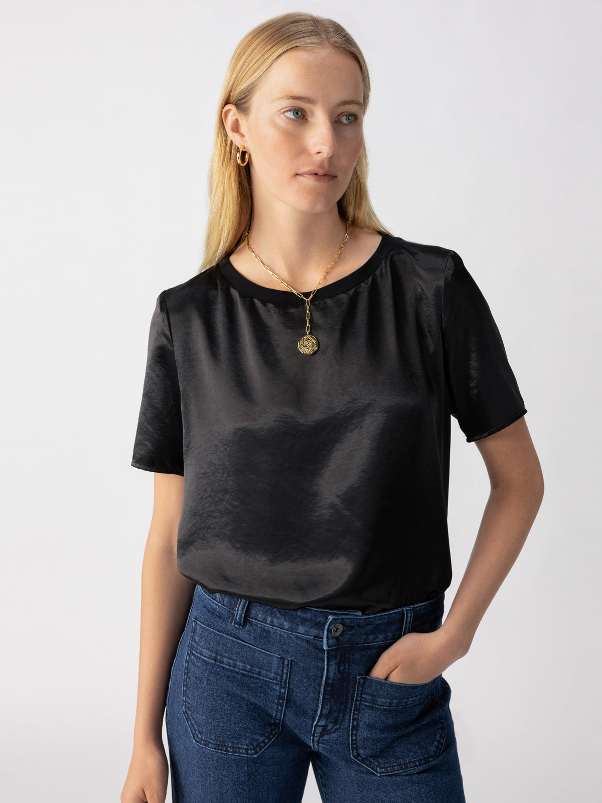 A person with long blond hair is wearing the Timeless Satin Tee Black from Sanctuary Clothing, accompanied by blue jeans and a gold necklace with a pendant. They have one hand in their pocket and are looking away from the camera against a plain, light-colored background.