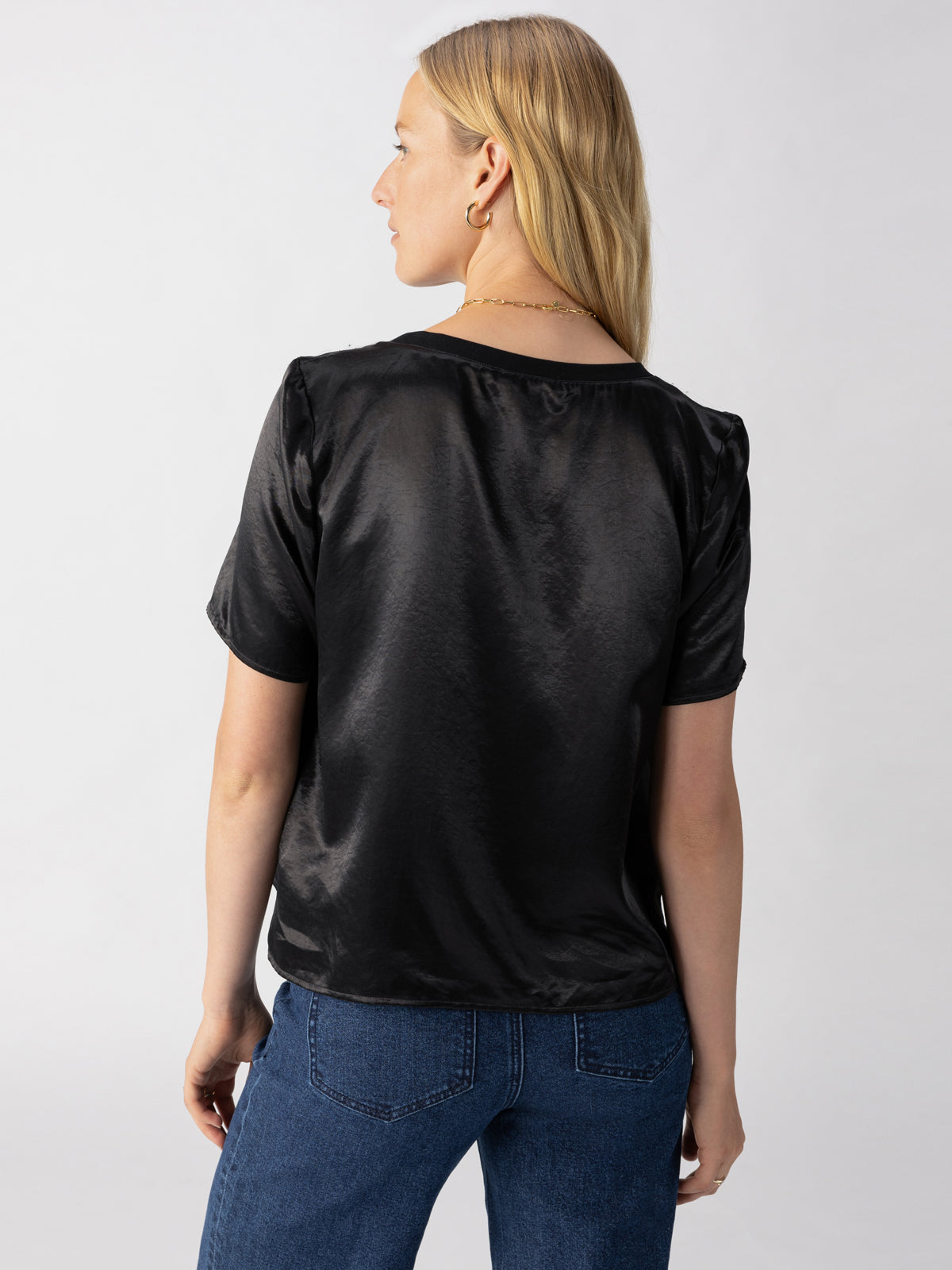 A woman with long blonde hair, seen from behind, is wearing a short-sleeved Timeless Satin Tee Black by Sanctuary Clothing and blue jeans. She has a gold necklace and hoop earrings against a plain, light-colored background.