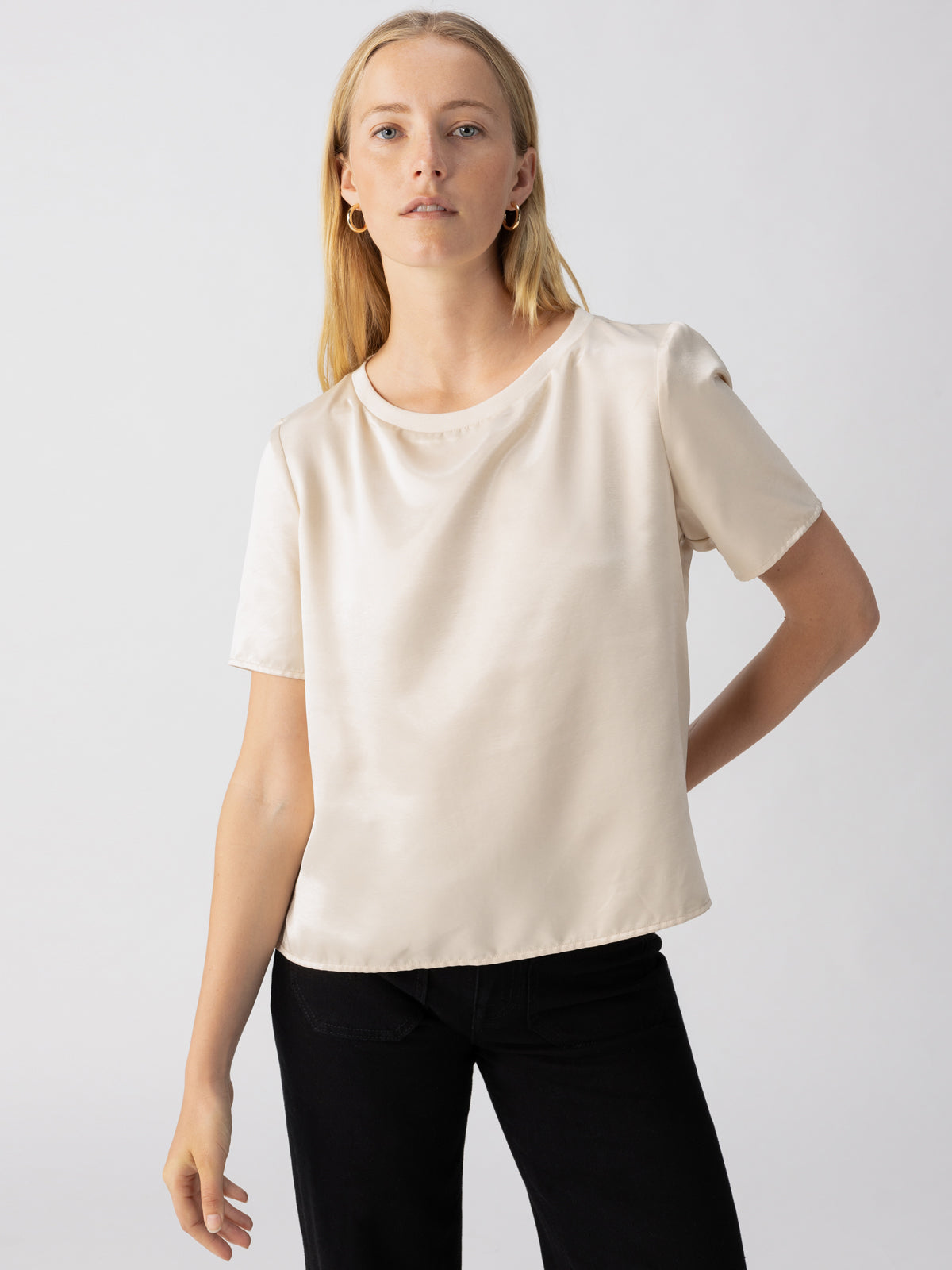 A person with long, light hair stands against a plain background, wearing the Timeless Satin Tee in Toasted Almond by Sanctuary Clothing and black pants. They have a relaxed expression and one hand placed on their hip.