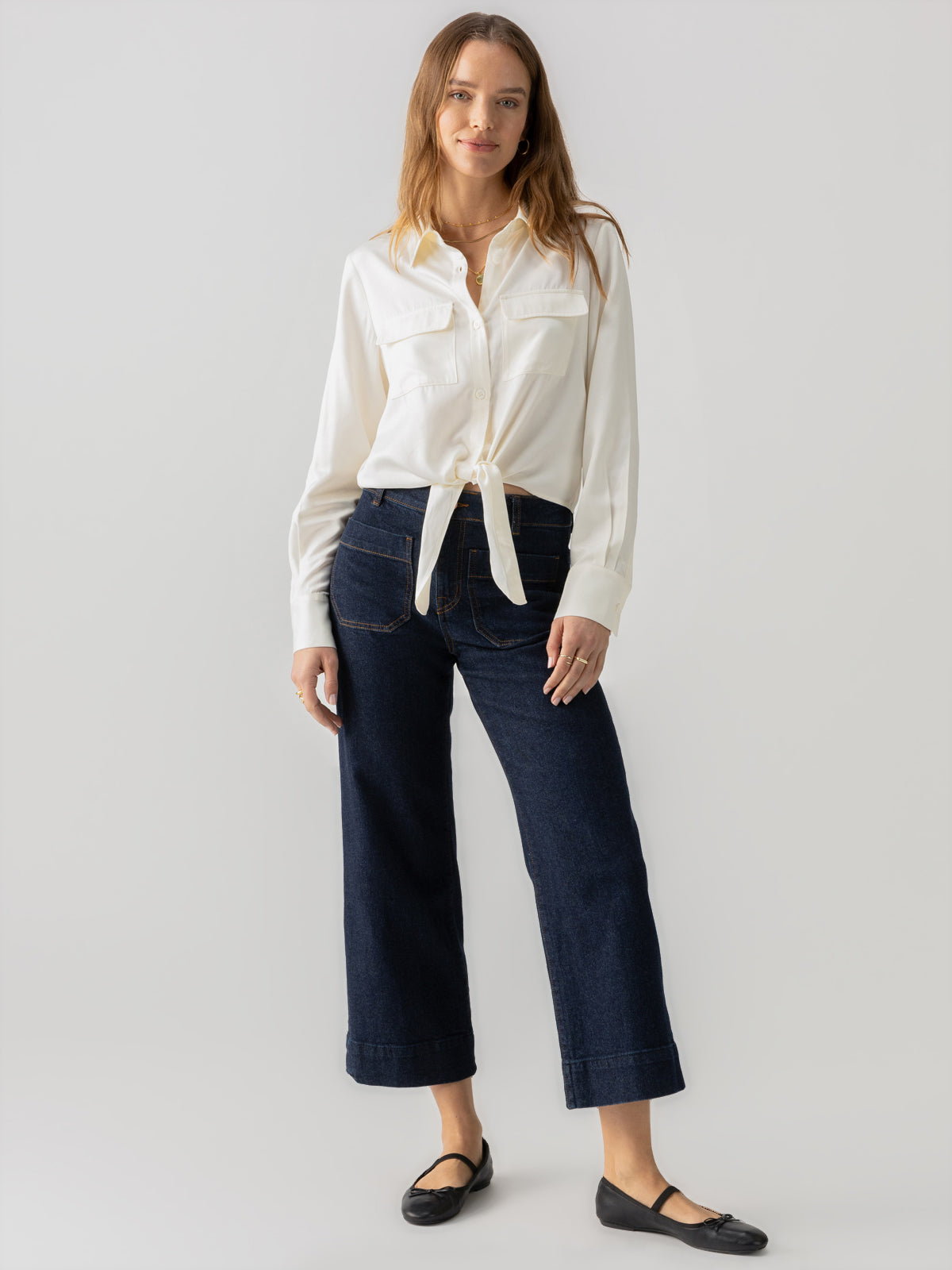 A woman with long hair is standing against a plain background. She is wearing the Lifetime Shirt Chalk by Sanctuary Clothing, tied at the waist, paired with dark high-waisted wide-leg jeans and black flat shoes. She has a relaxed posture and a neutral expression.