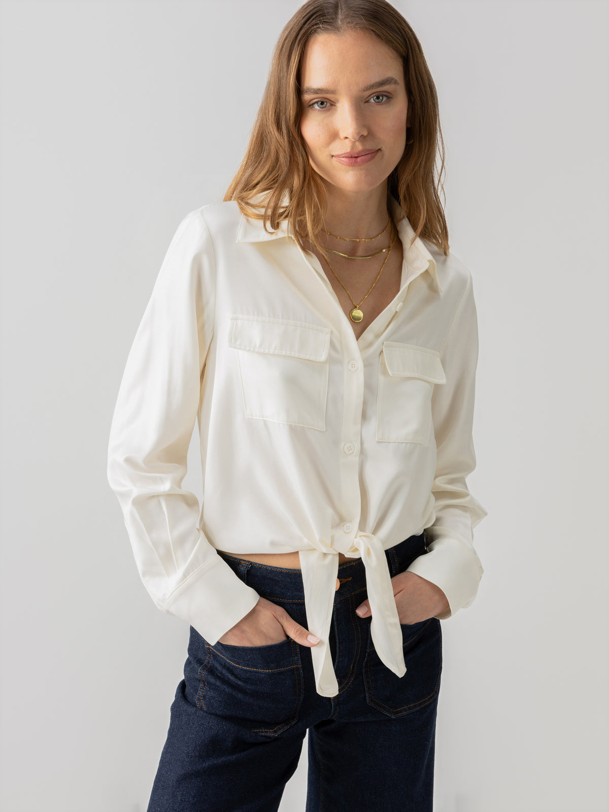 A person with long hair stands against a plain background, wearing the Sanctuary Clothing Lifetime Shirt Chalk tied at the waist and dark blue jeans. They have their hands in their pockets and are accessorized with layered necklaces. The expression on their face is calm and neutral.