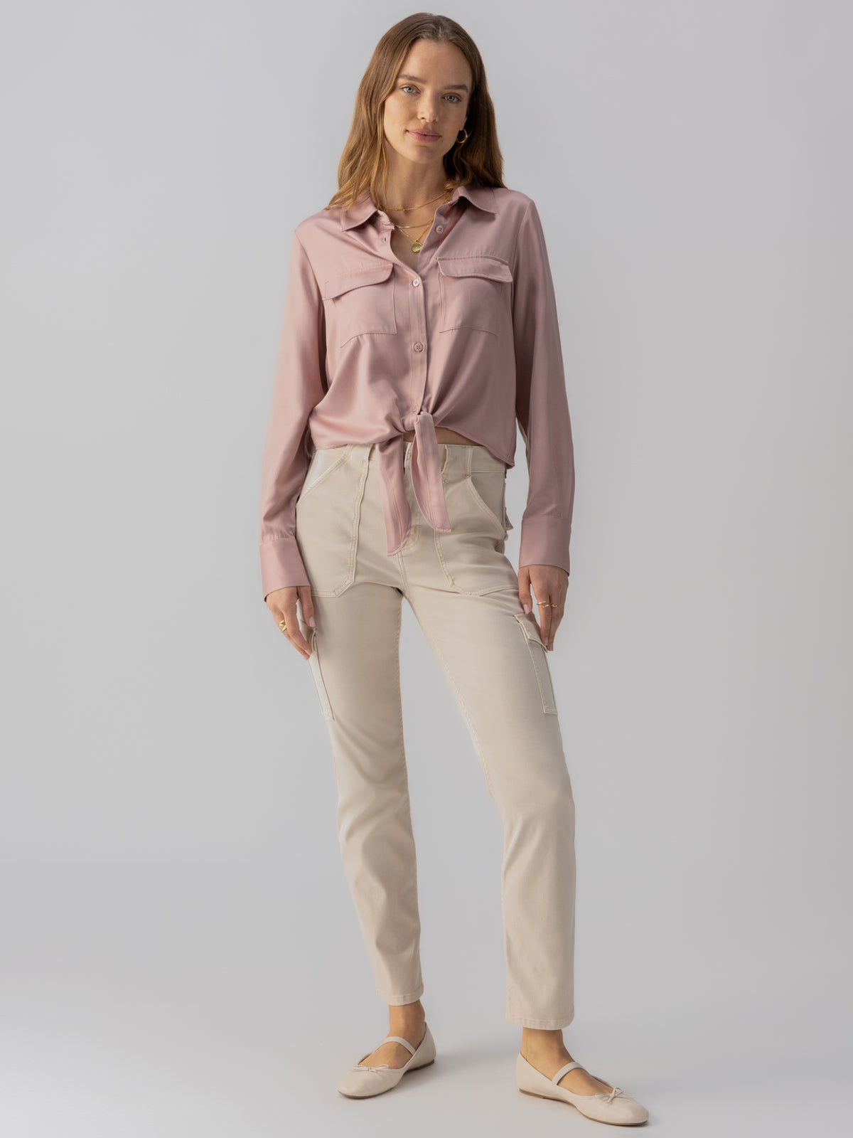 A woman with long hair stands against a light gray background. She is wearing the Lifetime Shirt in Smokey Rose from Sanctuary Clothing, a long-sleeve blouse tied at the waist, paired with light beige pants. She completes her outfit with beige flats, posing with a relaxed, soft smile.