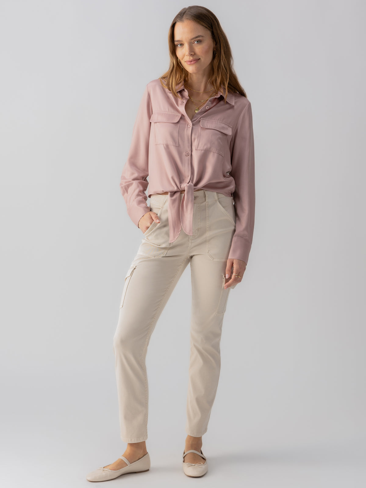 A woman stands against a light grey background, wearing a dusty pink button-up shirt with front pockets and a tie at the waist. She is paired with beige casual pants, specifically the Sculpted Hayden Cargo in Toasted Almond by Sanctuary Clothing. With her hands in her pockets, she completes her look with light beige flats and has long, wavy hair cascading down.