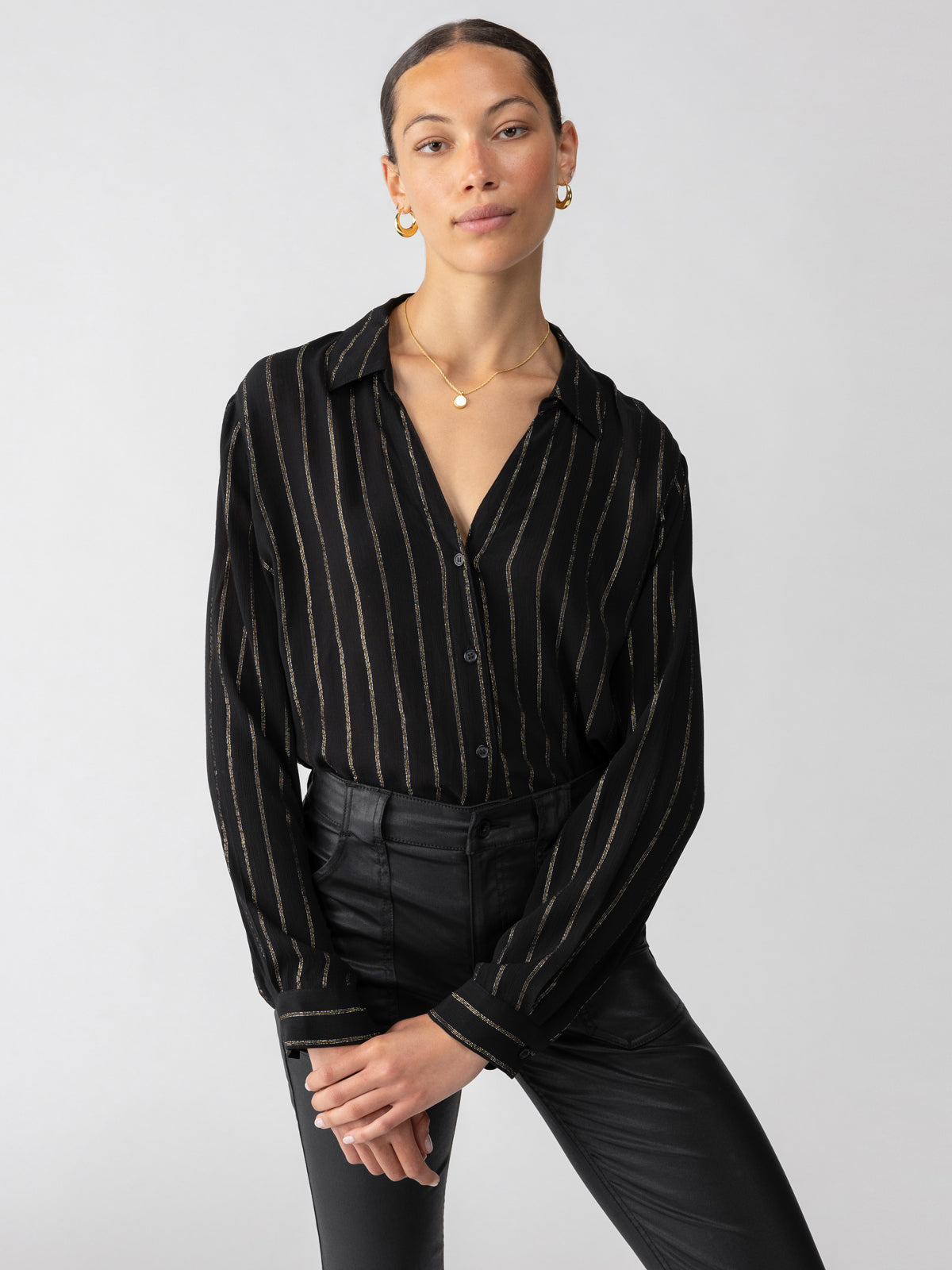 A person with pulled-back hair is wearing Sanctuary Clothing's Metallic Shirt Black, a long-sleeve button-up shirt with gold stripes, paired with black leather pants and gold hoop earrings. They are standing against a neutral background, hands gently clasped in front, and looking confidently at the camera.