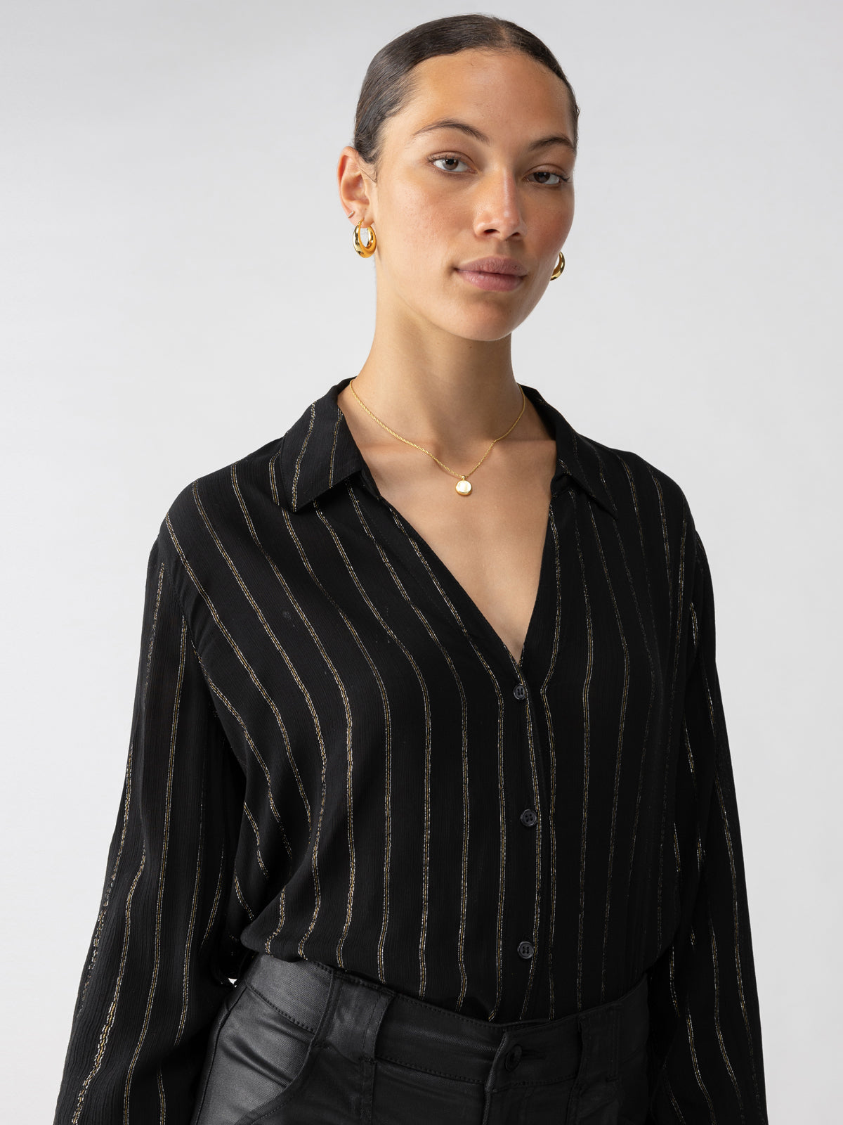 A person with a neat bun hairstyle poses confidently. They are wearing Sanctuary Clothing's Metallic Shirt Black, which is a black, long-sleeved shirt adorned with thin vertical golden stripes, paired with black pants. They accessorize with small hoop earrings and a delicate necklace featuring a round pendant against a plain white background.