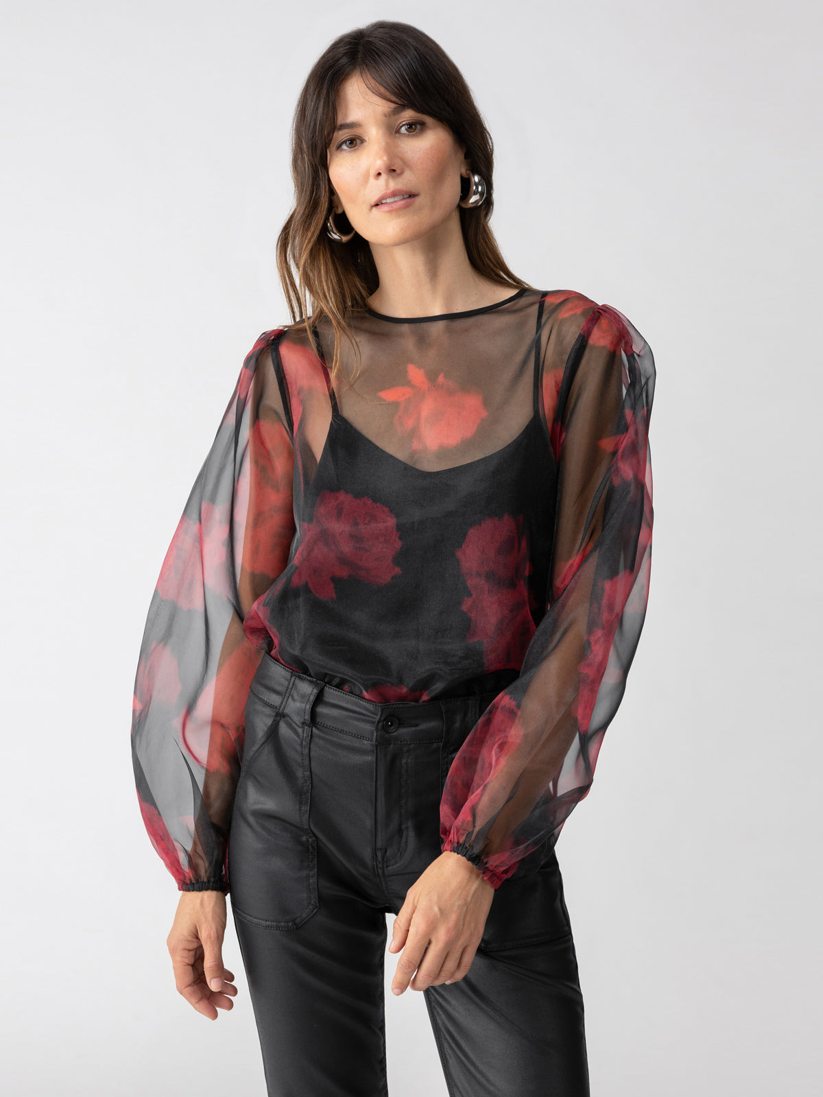 A woman with long brown hair is wearing the Moody Sheer Blouse Moody Rose by Sanctuary Clothing, featuring red floral patterns over a black camisole, paired with black leather pants. She stands against a plain light grey background and accessorizes with large hoop earrings.