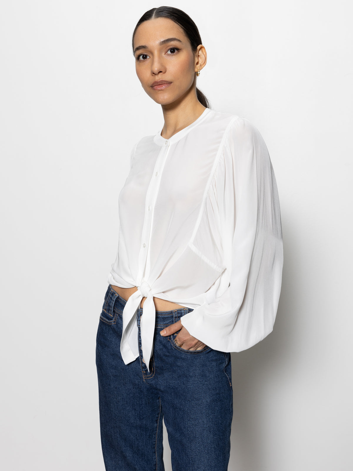 A person with long dark hair is wearing a tie front volume blouse in chalk by Sanctuary Clothing, paired with blue jeans. They are standing against a plain white background, looking slightly to their right with a neutral expression.