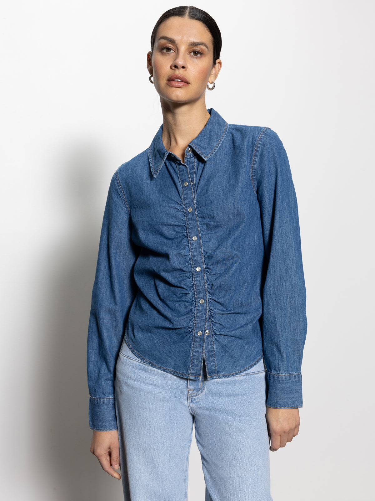 A person wearing Sanctuary Clothing's "denim dream girl top ball park" stands against a plain white background. They have pulled-back dark hair and are paired with light blue jeans.