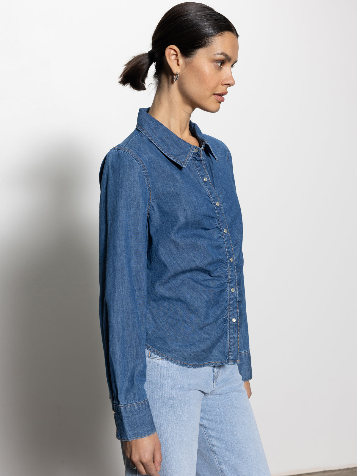 A woman in a Sanctuary Clothing "denim dream girl top ball park" and light blue jeans stands in profile against a plain background, her hair tied back.