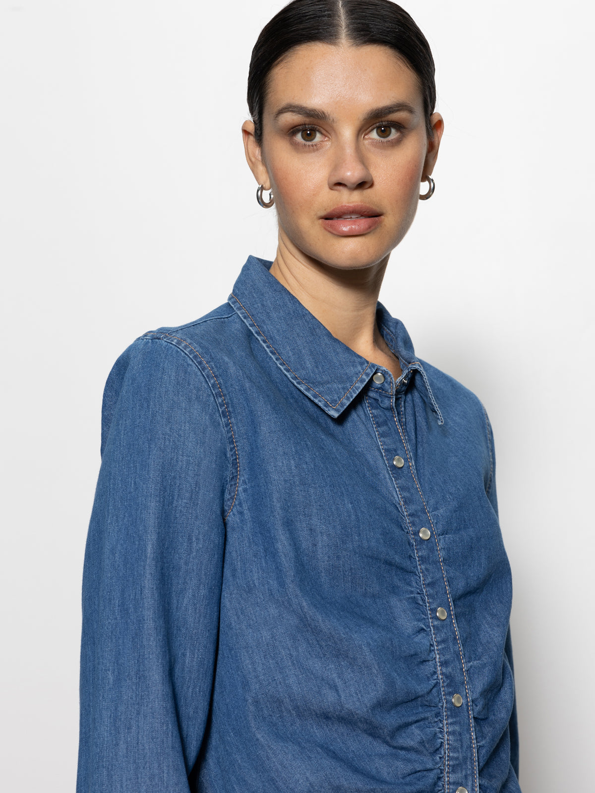 A person with dark hair tied back, wearing a Sanctuary Clothing denim dream girl top ball park and hoop earrings, is looking at the camera against a plain white background.