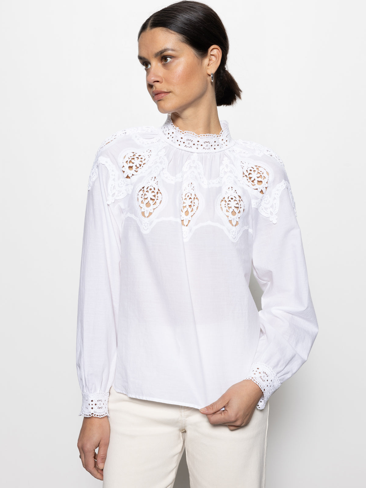 Wearing Sanctuary Clothing's lace popover blouse in white, featuring intricate lace detailing on the shoulders and chest, a person stands against a plain background. They have dark hair pulled back, look to the side, and rest their hand on their hip.