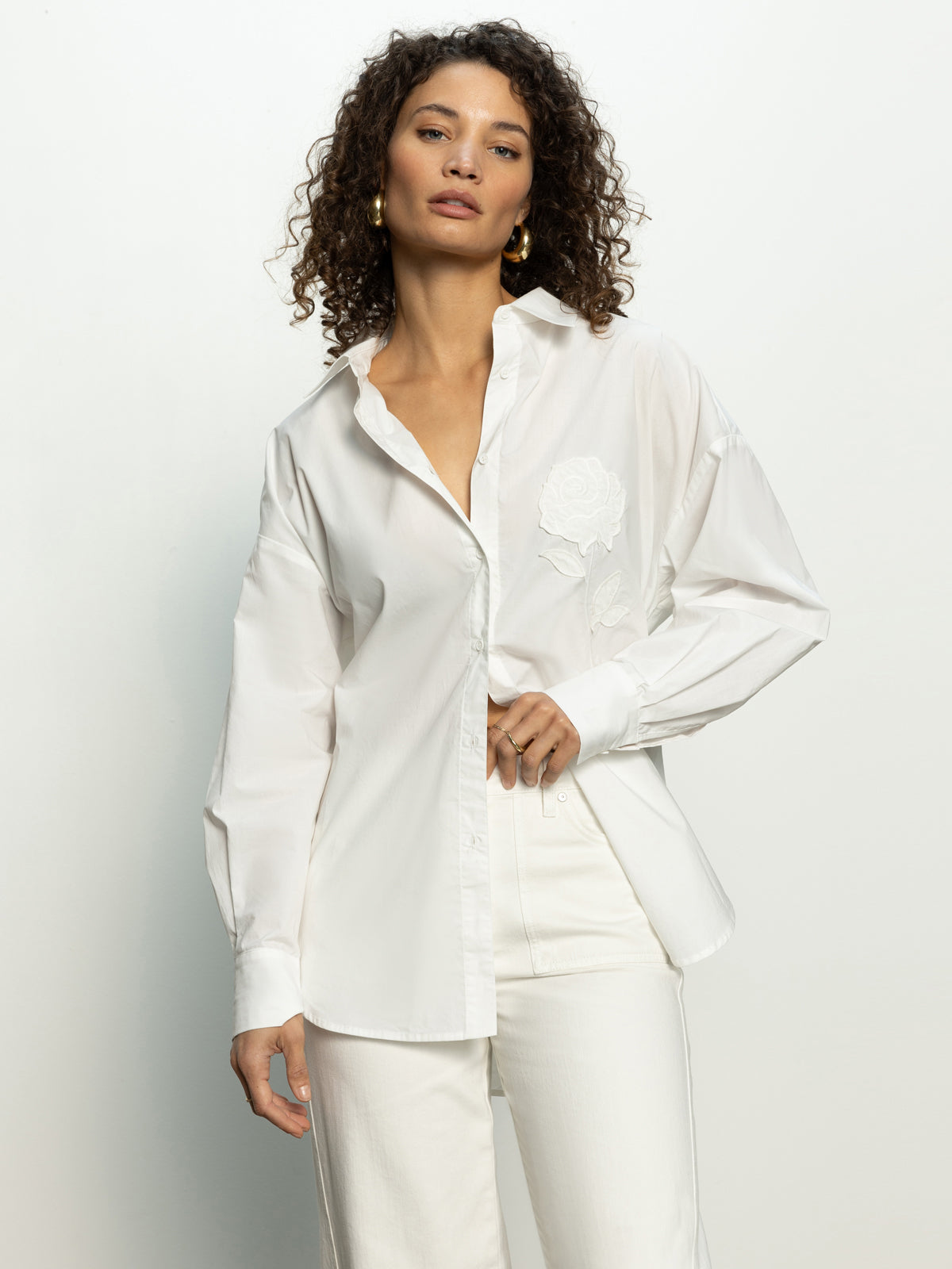 A person with curly hair models Sanctuary Clothing's "boyfriend tie back shirt white" against a plain background. They complete the look with white pants and gold hoop earrings, posing with a relaxed and confident expression.