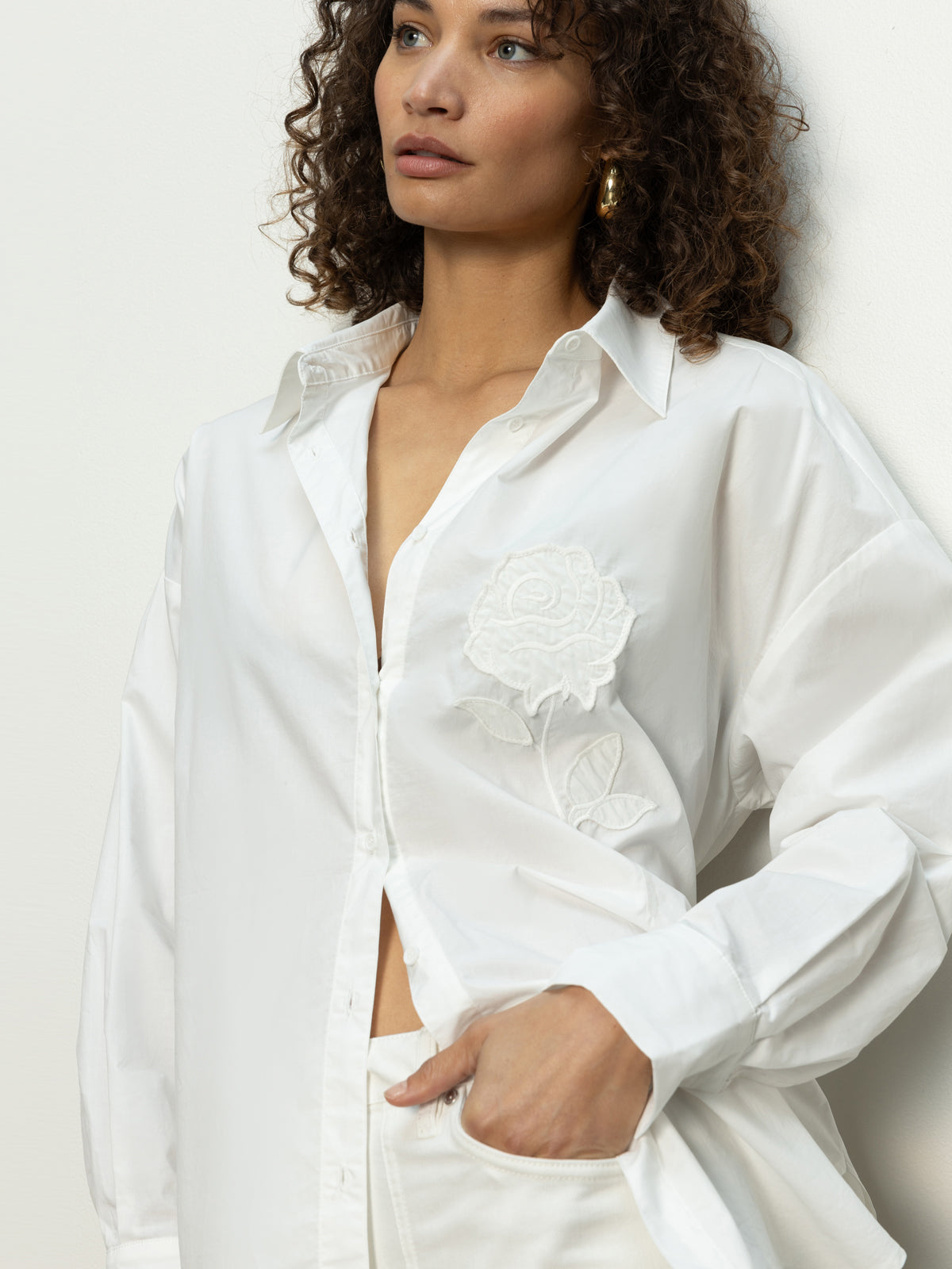 A woman with curly hair wears a partially unbuttoned Sanctuary Clothing "boyfriend tie back shirt" in white, revealing a white top underneath. The shirt has prominent floral embroidery. She casually looks away with one hand in her pocket.