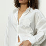 A woman with curly hair wears a partially unbuttoned Sanctuary Clothing "boyfriend tie back shirt" in white, revealing a white top underneath. The shirt has prominent floral embroidery. She casually looks away with one hand in her pocket.