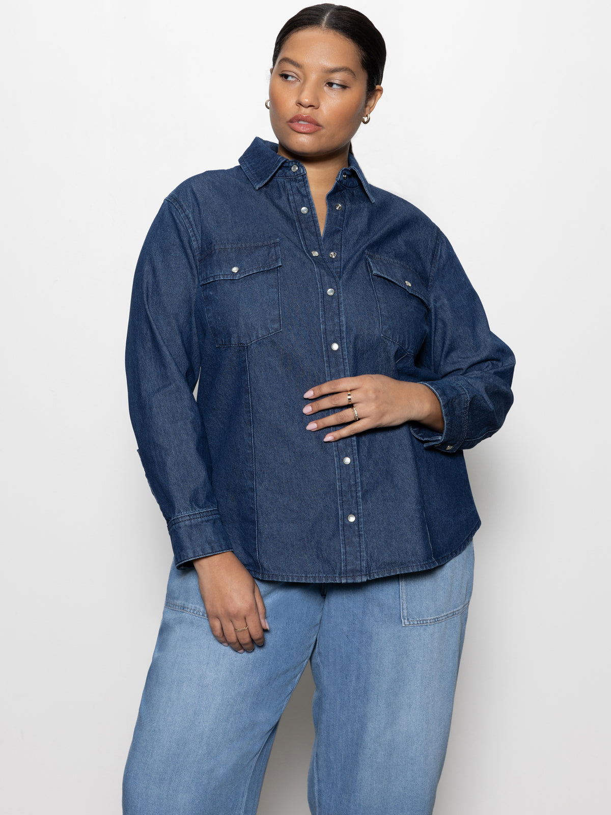 A person wearing the Sanctuary Clothing denim western shirt blue slate inclusive collection stands against a plain white background, with their right hand on their hip while looking slightly to the left, paired with light blue jeans.