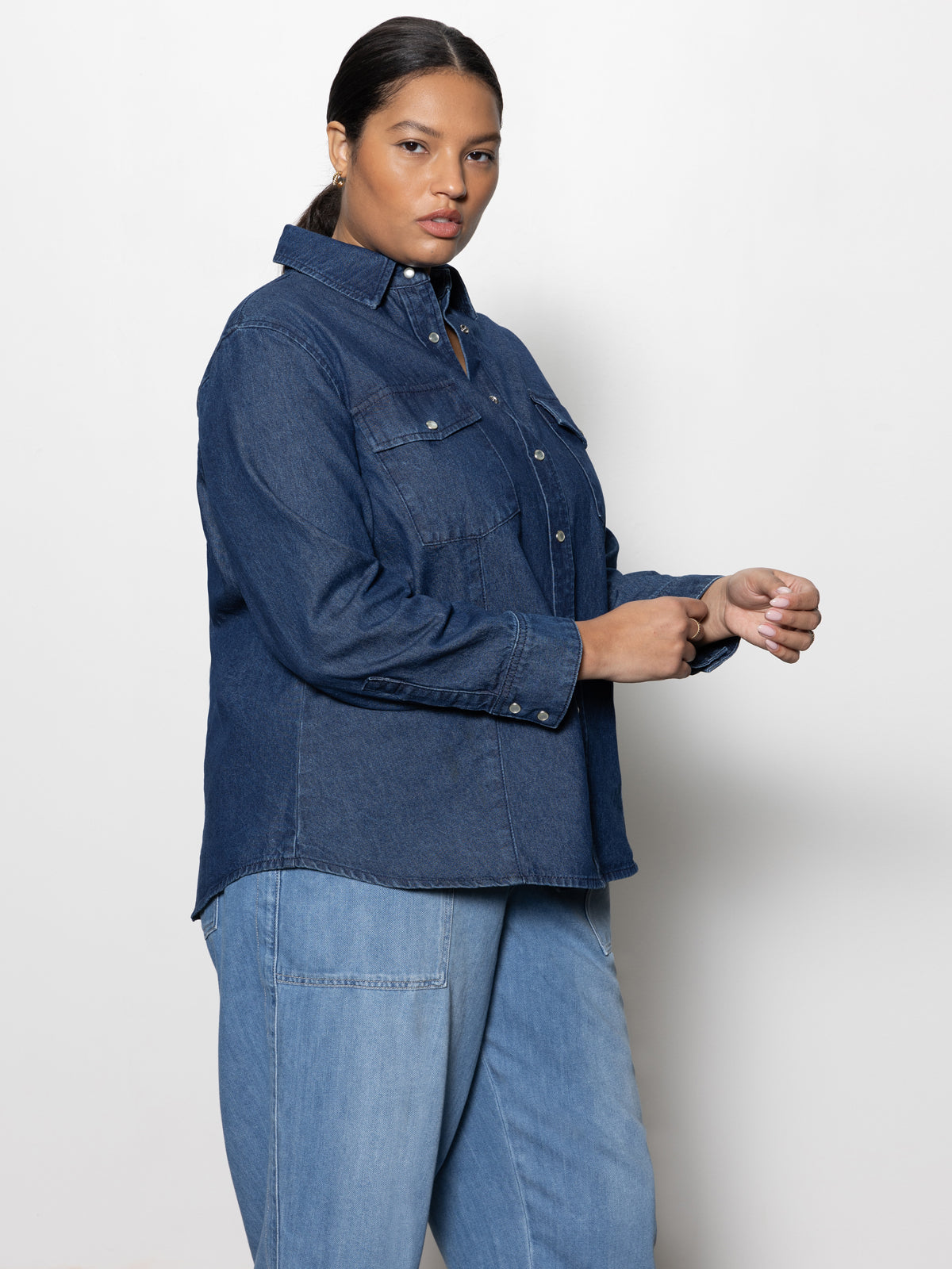 A woman in the Denim Western Shirt Blue Slate Inclusive Collection from Sanctuary Clothing stands against a white backdrop, looking to the side with a neutral expression while adjusting her sleeves. Her hair is tied back.