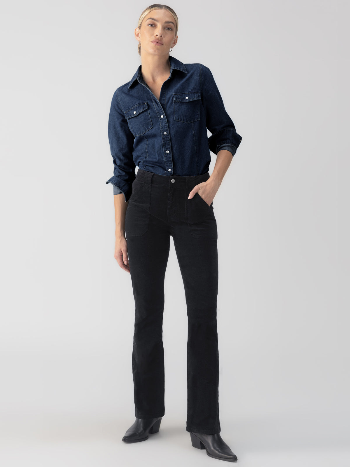 The individual stands confidently against a plain, light-colored background wearing Sanctuary Clothing's "The Denim Western Shirt Blue Slate." They have tucked the shirt into dark, high-waisted pants and completed the look with black boots, their hair neatly tied back.