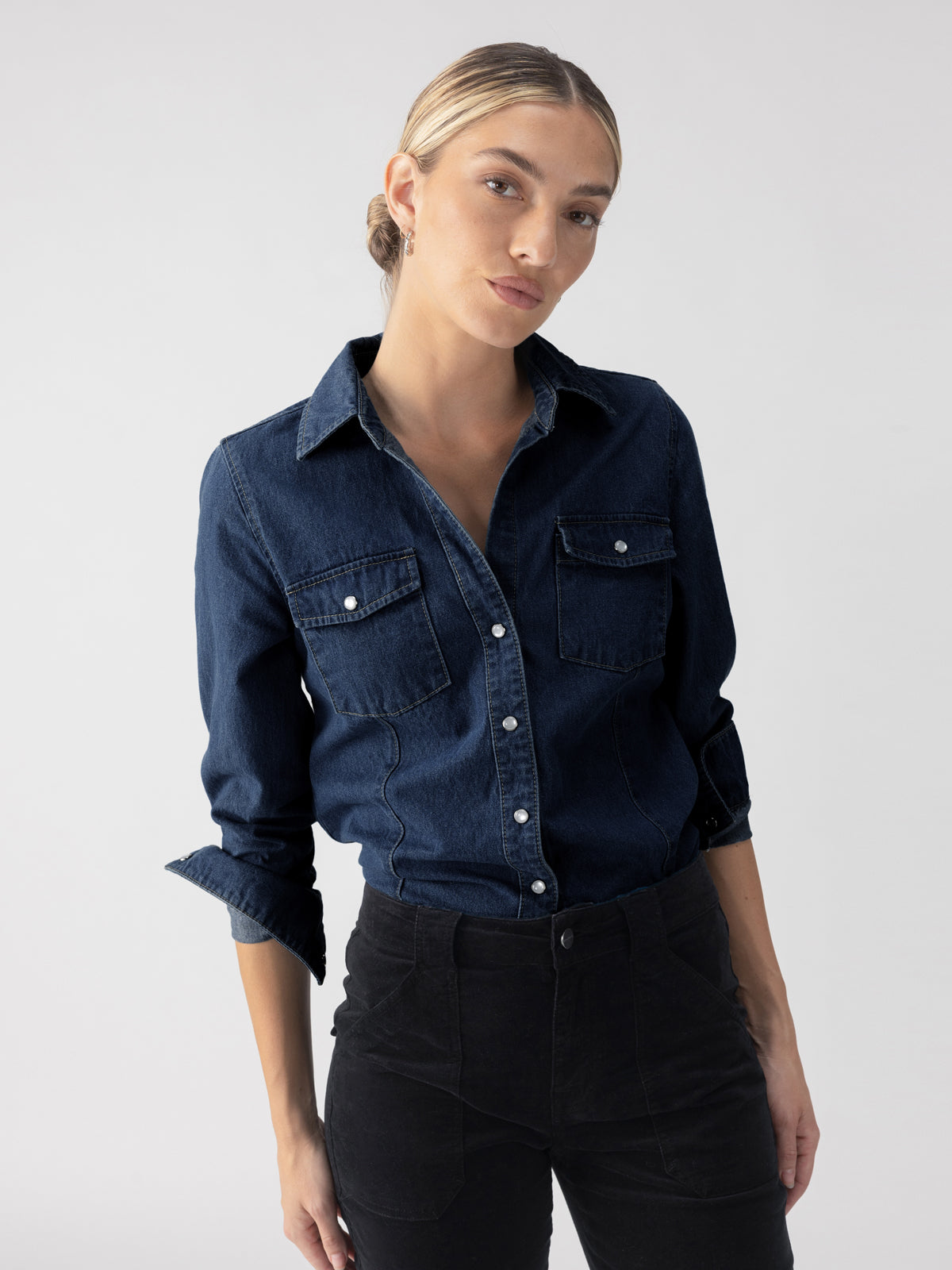 A person with light brown hair pulled back stands against a plain background, wearing the Denim Western Shirt Blue Slate from Sanctuary Clothing and black pants. The person's expression is neutral, and their posture is relaxed with their hands in their pockets.