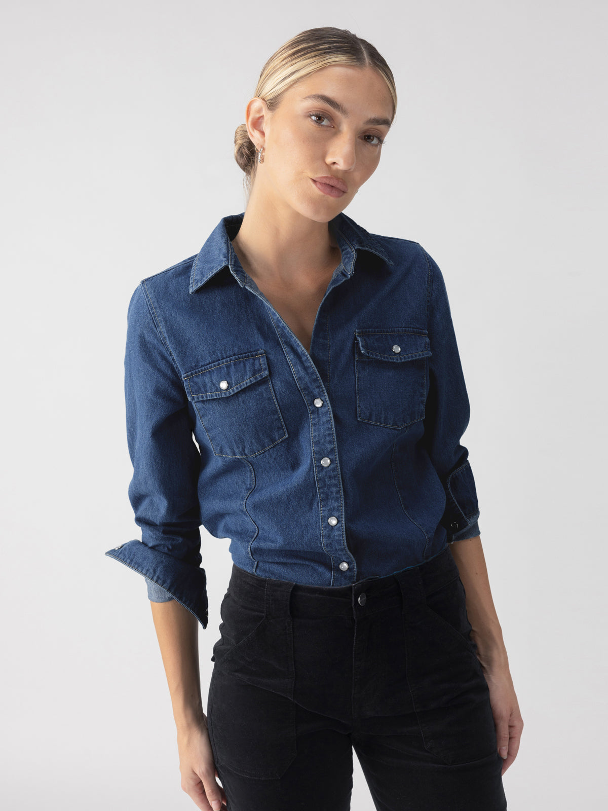 A person wearing The Denim Western Shirt Blue Slate by Sanctuary Clothing with rolled-up sleeves and black pants stands against a plain background. Their hair is neatly tied back, and they have a confident expression.