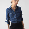 A person wearing The Denim Western Shirt Blue Slate by Sanctuary Clothing with rolled-up sleeves and black pants stands against a plain background. Their hair is neatly tied back, and they have a confident expression.