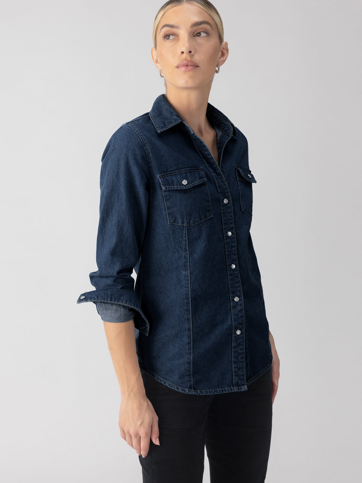 Person wearing "The Denim Western Shirt Blue Slate" by Sanctuary Clothing with rolled sleeves and black pants, standing against a plain background.