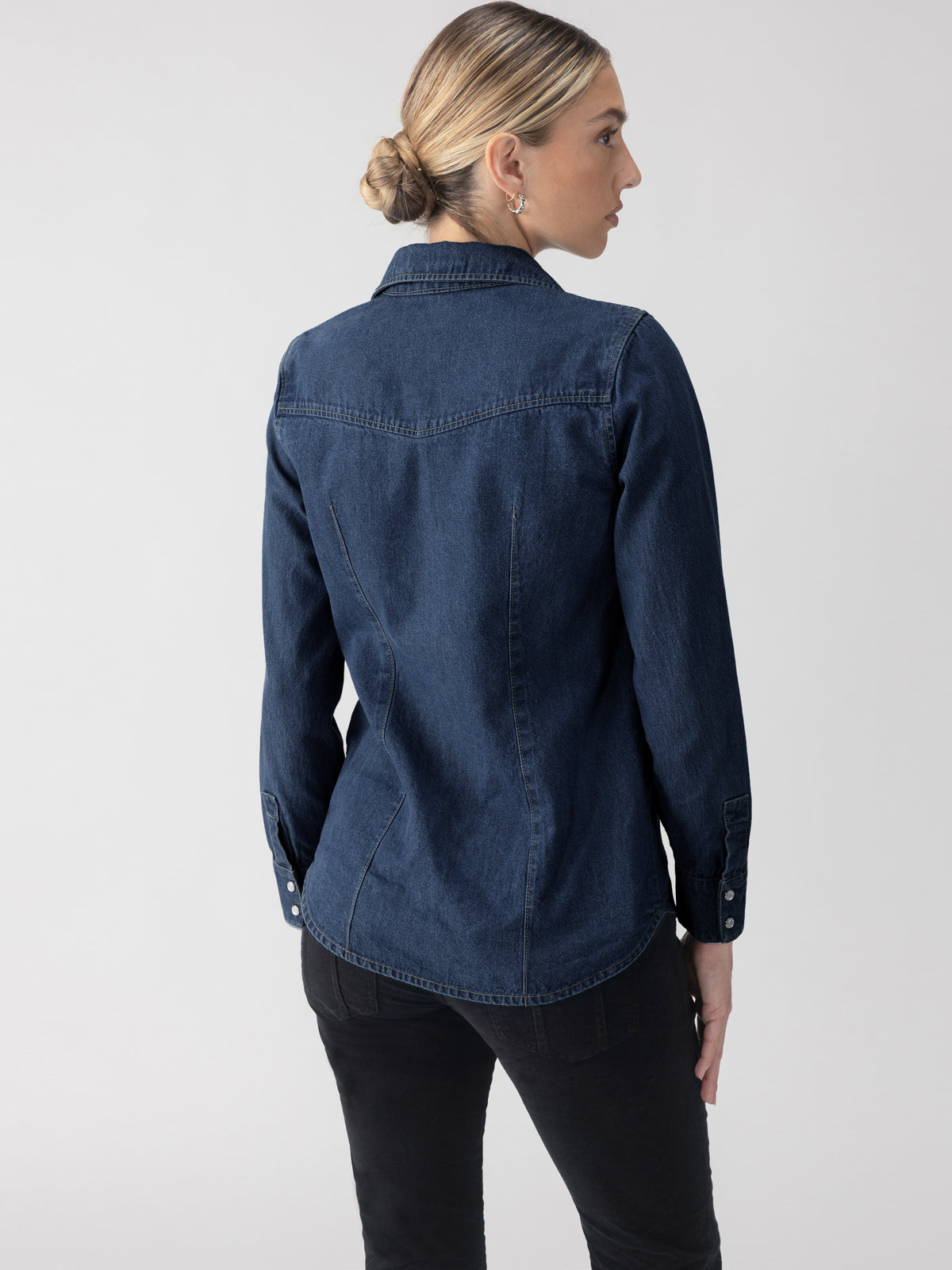 A person with blonde hair in a bun is wearing The Denim Western Shirt in Blue Slate by Sanctuary Clothing and black pants. They are standing and facing away from the camera, gazing to the side against a plain light gray background.