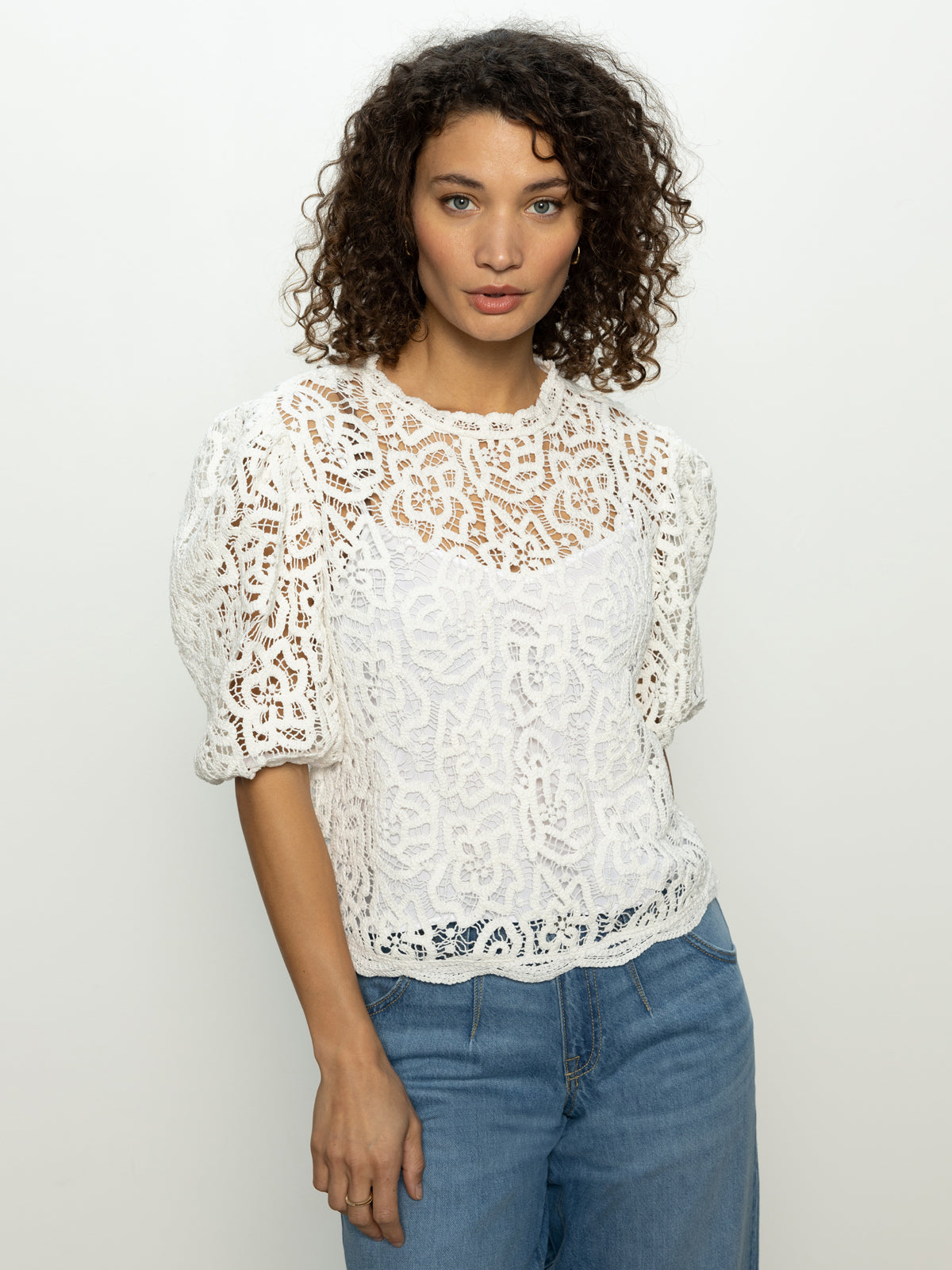 A person with curly hair is wearing the Cherished Lace Tee in white from Sanctuary Clothing, paired with blue jeans, standing against a plain, light background.