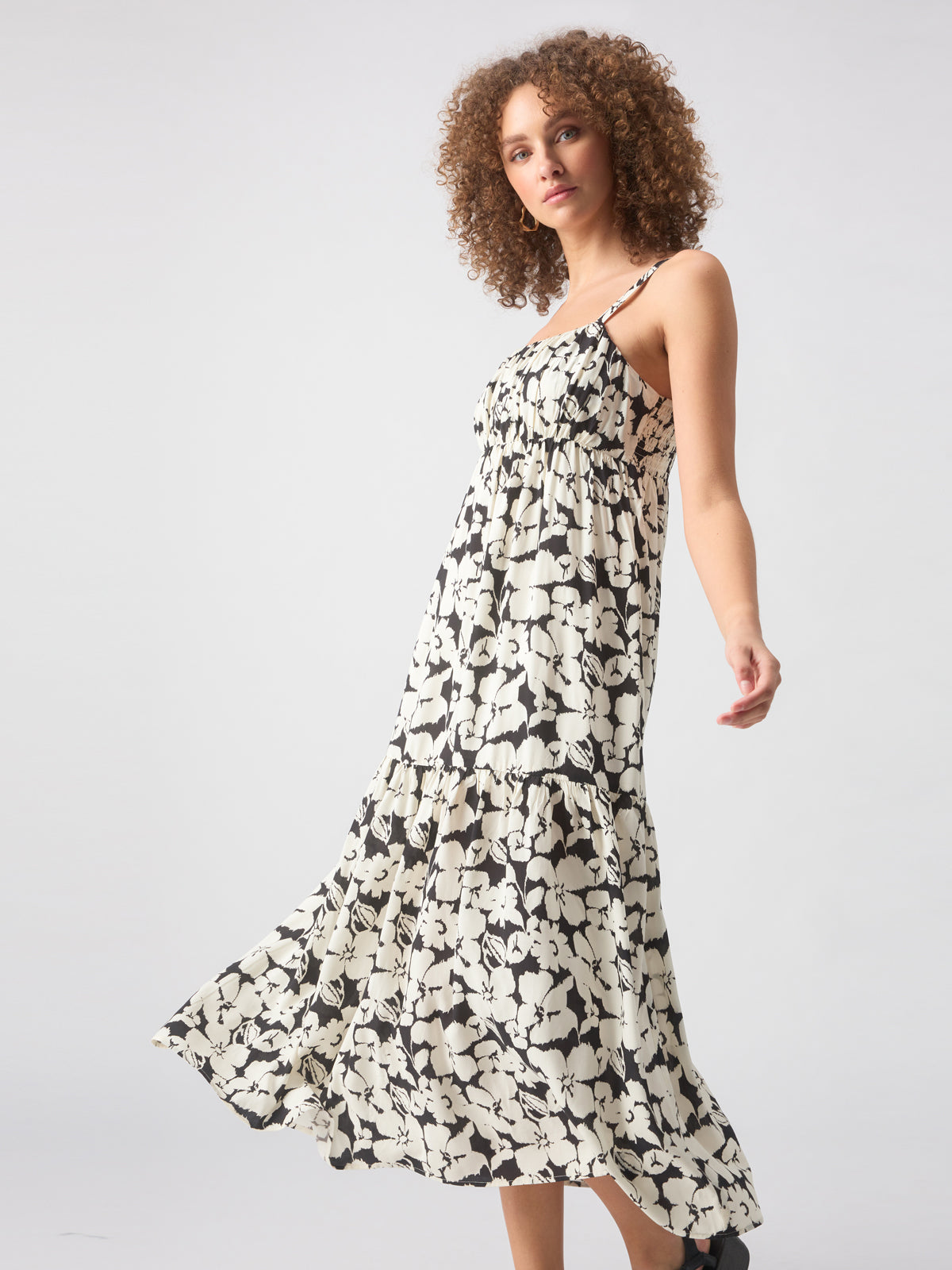 A woman with curly hair wears the Dropped Seam Maxi Echo Blooms, a long, sleeveless black and white floral dress by Sanctuary Clothing. She poses with one hand lifted, looking confident and elegant. The background is plain and light-colored, emphasizing her dress.