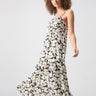 A woman with curly hair wears the Dropped Seam Maxi Echo Blooms, a long, sleeveless black and white floral dress by Sanctuary Clothing. She poses with one hand lifted, looking confident and elegant. The background is plain and light-colored, emphasizing her dress.