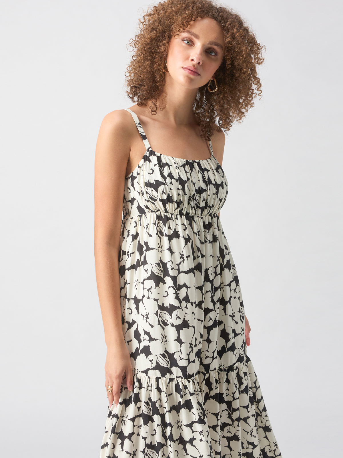 A woman with curly hair is wearing the Dropped Seam Maxi Echo Blooms dress by Sanctuary Clothing, a sleeveless, black and white floral maxi dress featuring a square neckline and a tiered skirt. She stands against a plain, light background, looking slightly down with a faint smile.