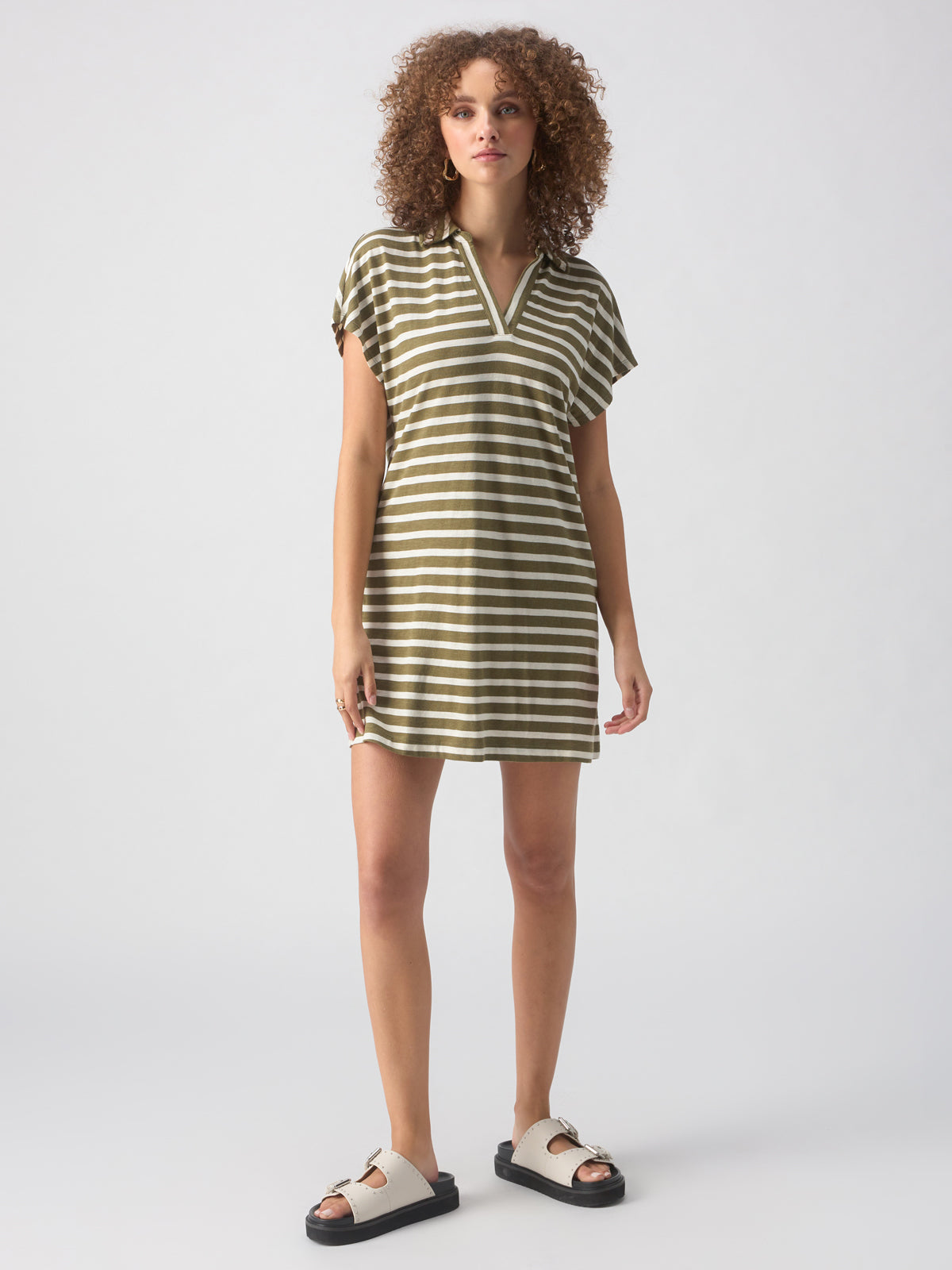 A person with curly hair stands against a plain background, wearing the Johnny Collar Tshirt Dress Light Ecru Olive Stripe from Sanctuary Clothing. The short-sleeved dress falls above the knees, and they are also wearing white slides with black soles.