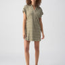 A person with curly hair stands against a plain background, wearing the Johnny Collar Tshirt Dress Light Ecru Olive Stripe from Sanctuary Clothing. The short-sleeved dress falls above the knees, and they are also wearing white slides with black soles.