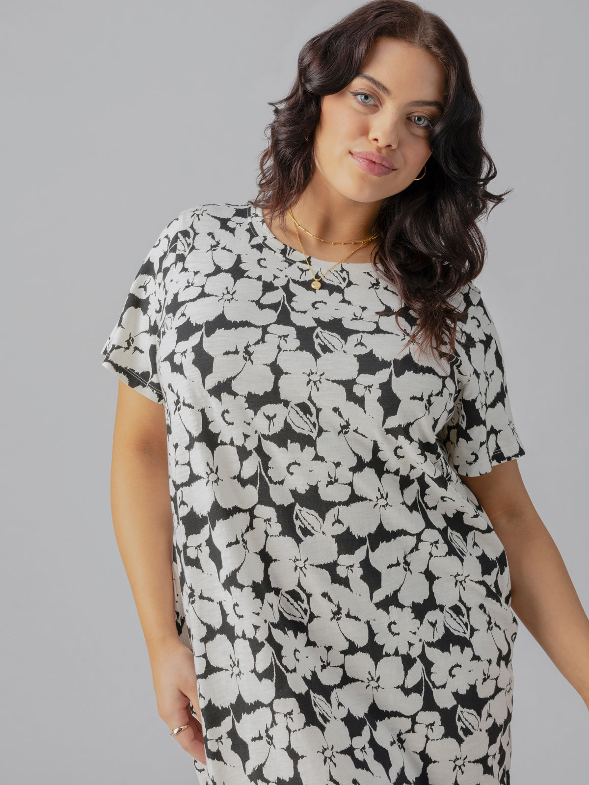 A woman with long, wavy dark hair is wearing The Only One T Shirt Dress Echo Blooms from Sanctuary Clothing's Inclusive Collection. She is standing against a plain grey background, looking at the camera with a slight smile, and has her left hand resting casually on her hip.