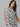 A woman with long, wavy dark hair is wearing The Only One T Shirt Dress Echo Blooms from Sanctuary Clothing's Inclusive Collection. She is standing against a plain grey background, looking at the camera with a slight smile, and has her left hand resting casually on her hip.