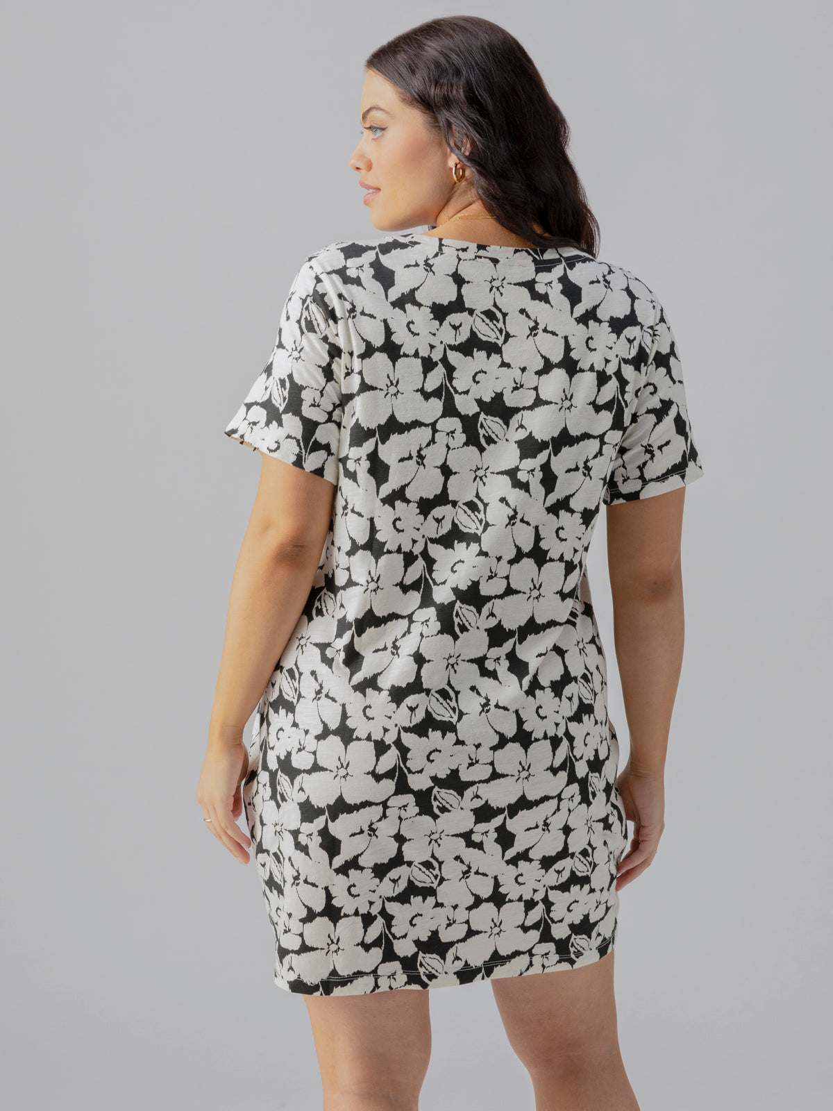 A woman with wavy hair is shown from the back, wearing The Only One T Shirt Dress Echo Blooms Inclusive Collection by Sanctuary Clothing. The dress features a short-sleeved, black and white floral pattern. She stands against a neutral gray background, with her hands resting at her sides.