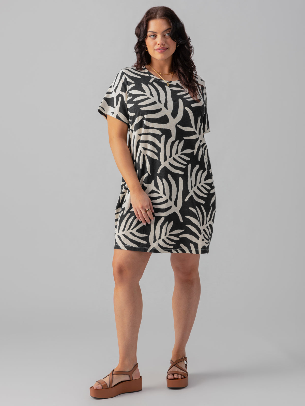 A person with long wavy hair is standing against a plain background, wearing The Only One T Shirt Dress Night Palm from the Inclusive Collection by Sanctuary Clothing, featuring a bold black and white leaf pattern. They are also sporting brown platform sandals. With a neutral expression, their arms hang relaxed at their sides.