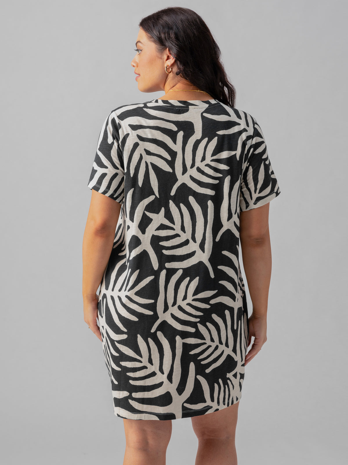 A woman with dark hair stands facing away from the camera, wearing The Only One T Shirt Dress Night Palm Inclusive Collection by Sanctuary Clothing. This short-sleeved, knee-length dress showcases a striking black and beige palm leaf pattern against a plain grey background.