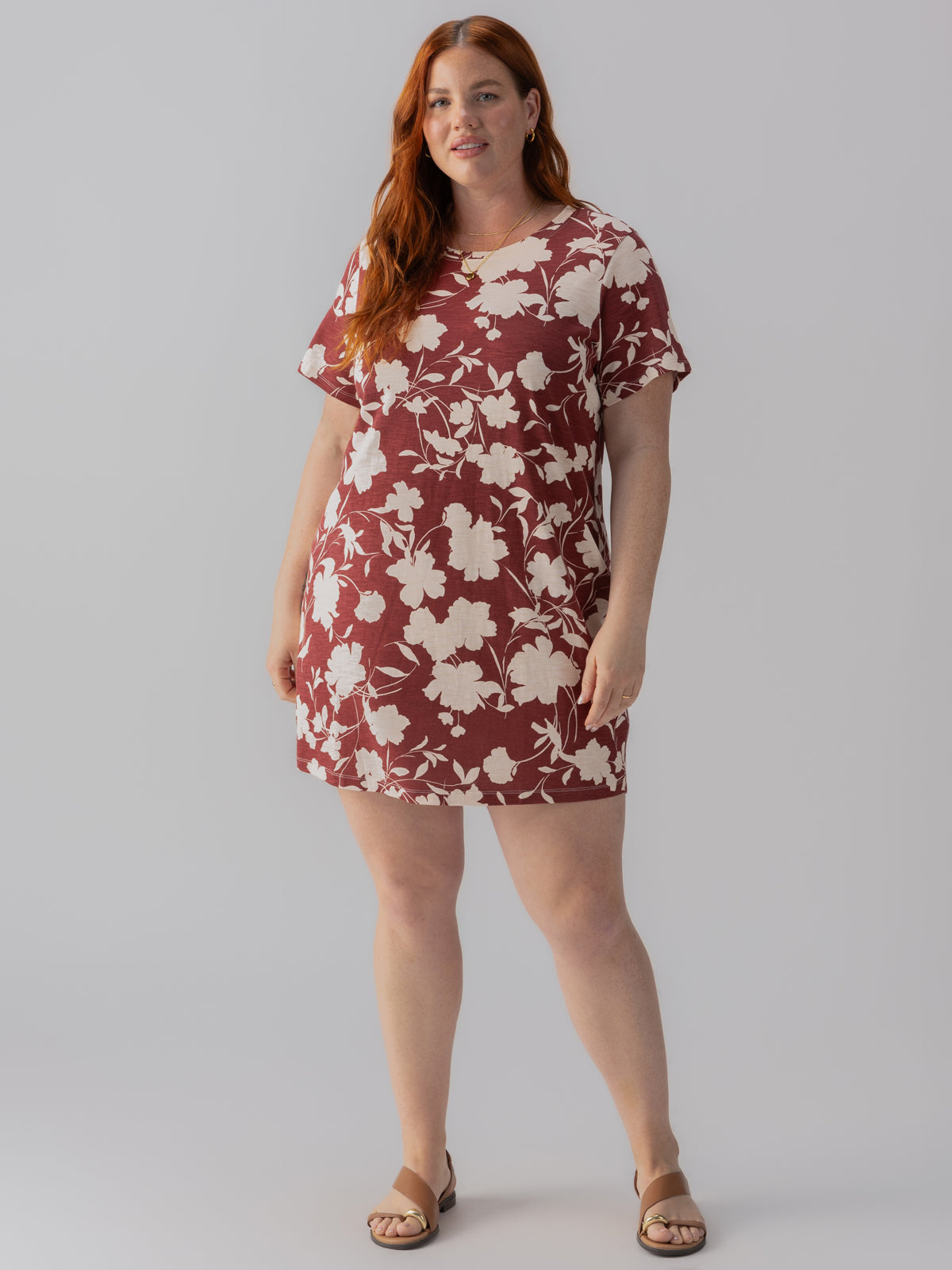 A woman with red hair is standing against a plain background. She is wearing "The Only One T Shirt Dress Warm Vista Inclusive Collection" by Sanctuary Clothing, which features a short, burgundy design with a large white floral pattern. She is also wearing tan sandals and smiling slightly.