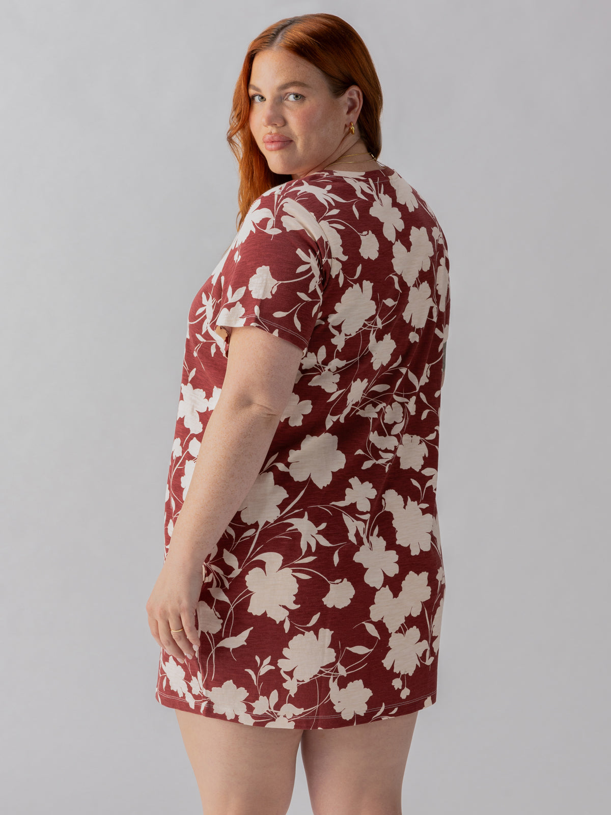 A woman with long red hair is wearing The Only One T Shirt Dress Warm Vista from the Inclusive Collection by Sanctuary Clothing. The dress features short sleeves and a maroon color adorned with a white floral pattern. She is standing sideways, slightly turning her head towards the camera against a plain, light gray background.