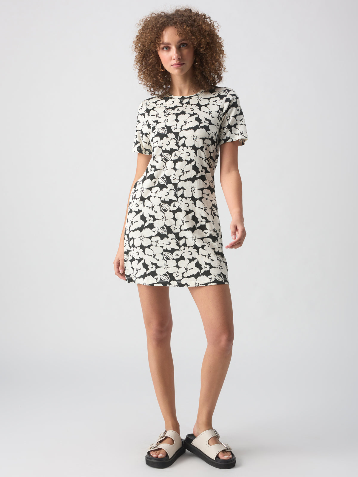 A person with curly hair stands against a plain grey background, wearing The Only One T Shirt Dress Echo Blooms by Sanctuary Clothing. They have on white sandals with black soles and buckles. The person maintains a neutral expression, with their arms relaxed at their sides.