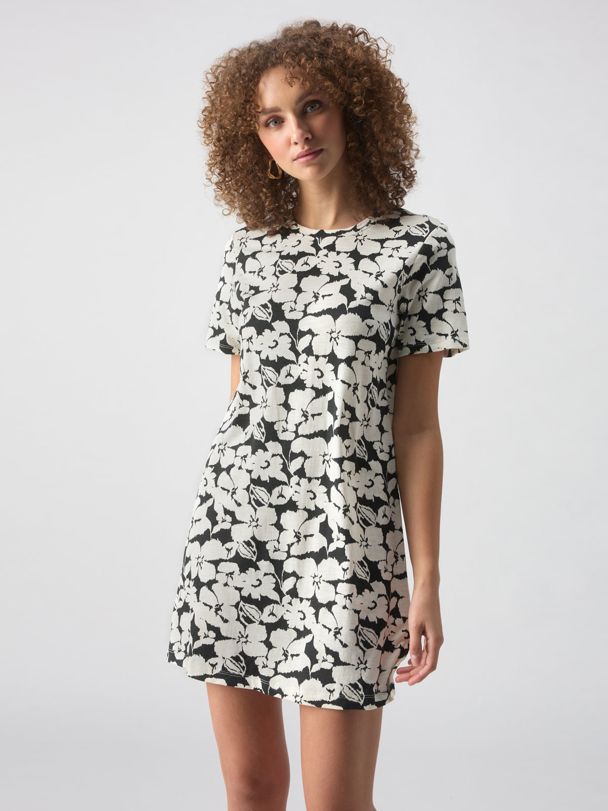 A person with curly hair is wearing The Only One T Shirt Dress Echo Blooms by Sanctuary Clothing. They are standing against a plain, light grey background, with a neutral expression on their face.