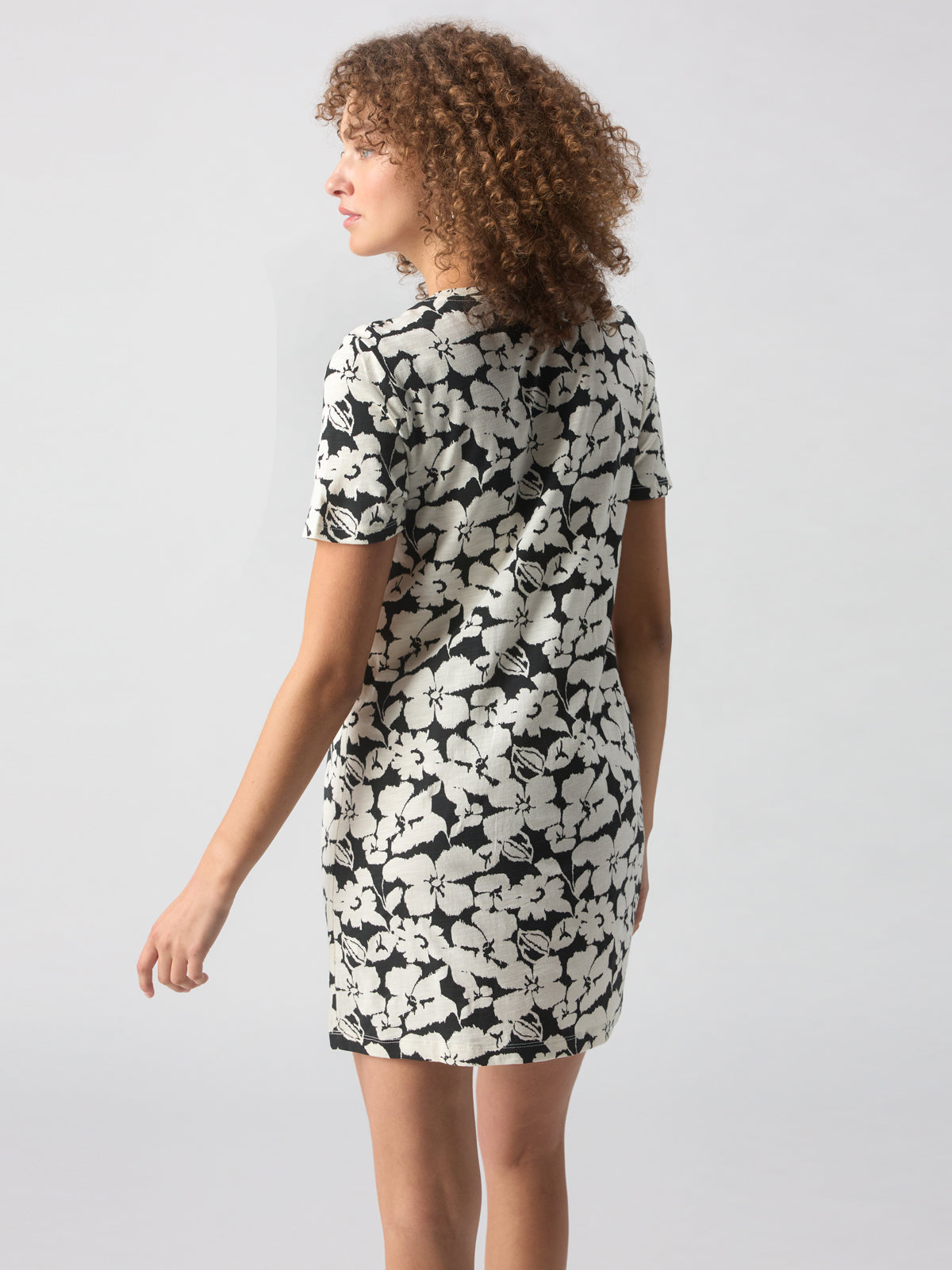 A person with curly hair is standing and facing away from the camera, wearing The Only One T Shirt Dress Echo Blooms by Sanctuary Clothing. The short-sleeved, black and white floral dress extends above the knees. Their right arm is slightly bent and positioned towards the side. The background is plain and light-colored.