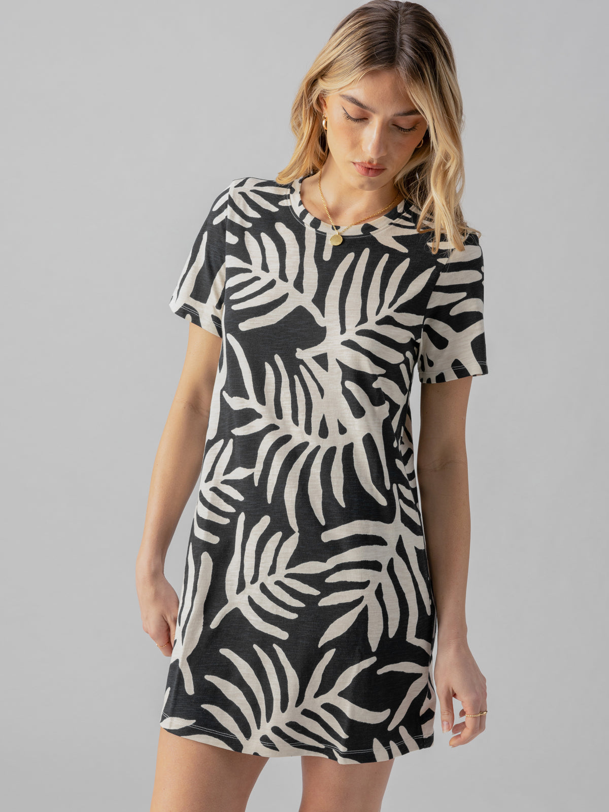 A person with wavy blonde hair is wearing The Only One T Shirt Dress Night Palm by Sanctuary Clothing, which features a short-sleeved, black and white leaf pattern. They are looking downwards with hands relaxed by their sides against a light gray background.