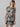 A person with wavy blonde hair is wearing The Only One T Shirt Dress Night Palm by Sanctuary Clothing, which features a short-sleeved, black and white leaf pattern. They are looking downwards with hands relaxed by their sides against a light gray background.