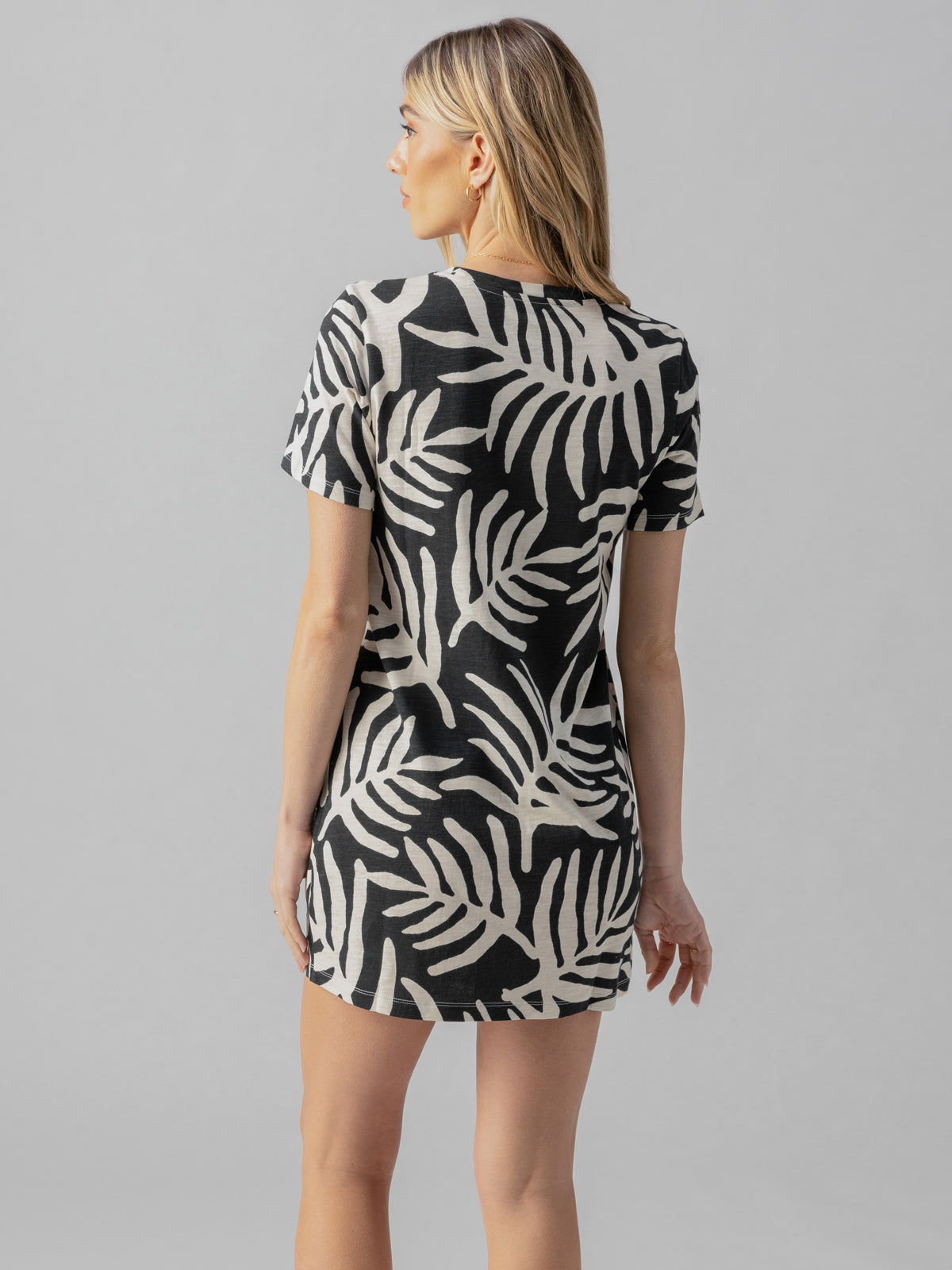 A person with long, light-colored hair is standing with their back to the camera. They are wearing The Only One T Shirt Dress Night Palm by Sanctuary Clothing, featuring a black and white leaf pattern. The background is plain and gray.