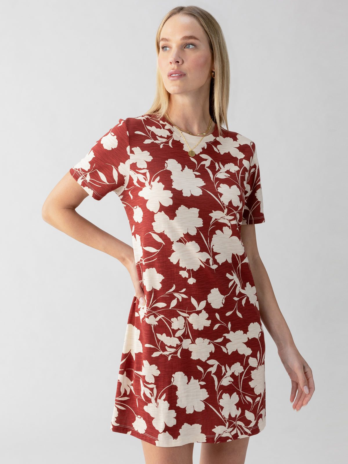 A woman with long blonde hair is modeling Sanctuary Clothing's The Only One T-Shirt Dress Warm Vista, featuring short sleeves and a white floral pattern on a red fabric. She is looking toward her left with her right hand on her hip against a plain, light-colored background.